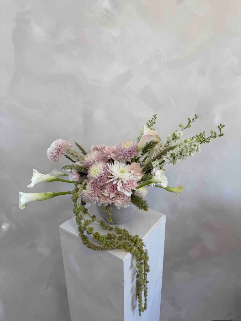 Soft pink and white arrangement with chrysanthemums, calla lilies, and trailing green amaranthus in a light gray bowl on a white pedestal