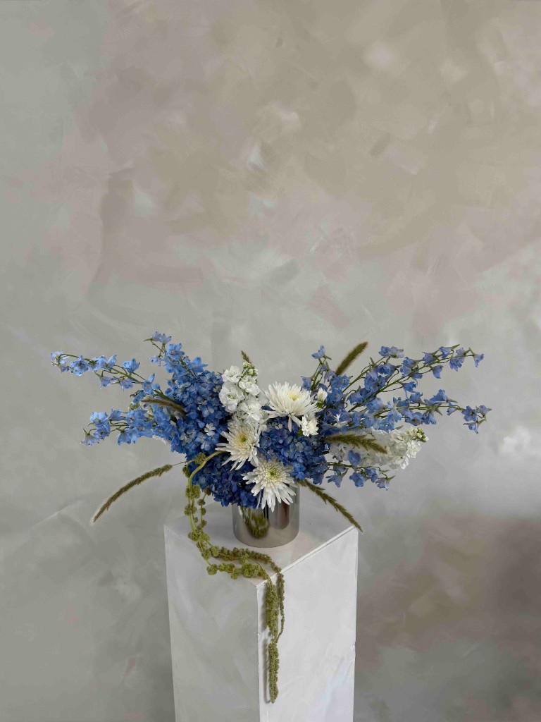 Wide blue and white arrangement with hydrangeas, delphiniums, white chrysanthemums, and trailing green amaranthus in a silver vase on a white pedestal