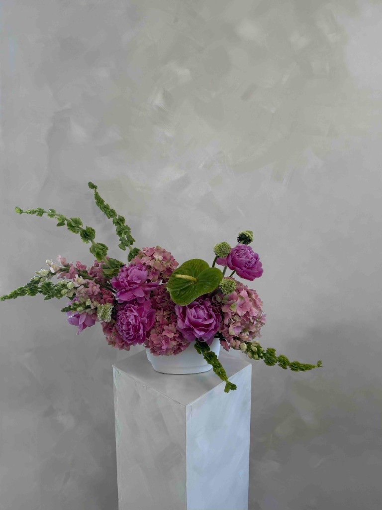 Bright pink and green arrangement with magenta peonies, dusty rose hydrangeas, green anthurium, and bells of Ireland in a white bowl on a pedestal