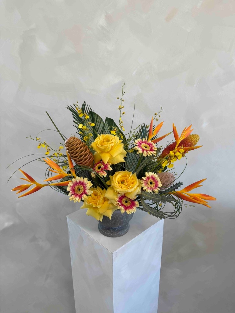 Whimsical tropical arrangement with yellow roses, gerbera daisies, heliconia, beehive ginger, and oncidium orchids in a dark gray vase on a white pedestal