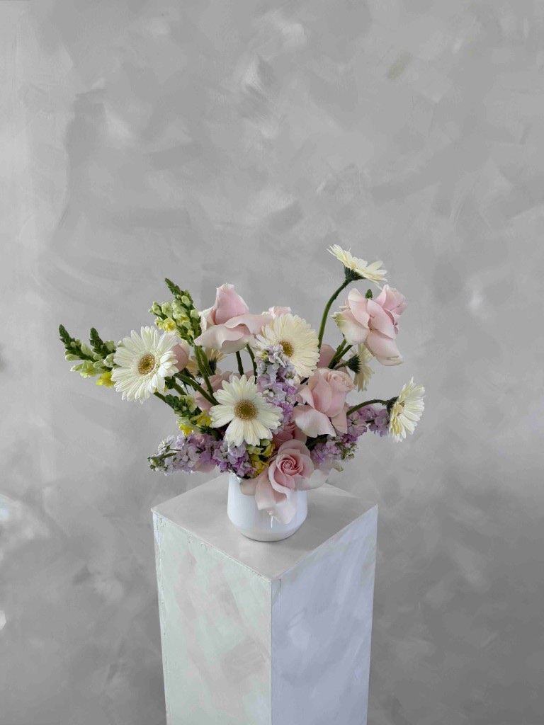 Pastel garden-style arrangement with light pink roses, white gerbera daisies, lavender stock, and green snapdragon buds in a white vase on a pedestal