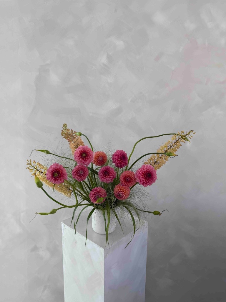 Whimsical arrangement with pink and orange dahlias, yellow flower spikes, and airy green stems in a white vase on a pedestal against a textured gray wall