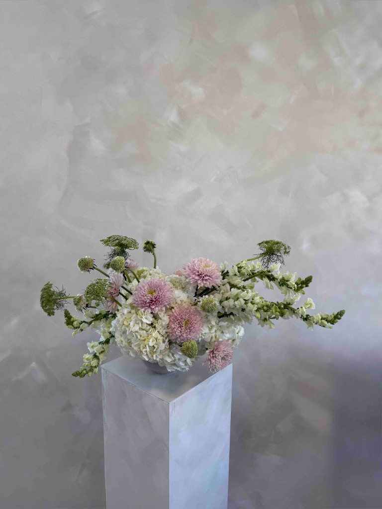 Pink, green, and white arrangement with light pink dahlias, white hydrangeas, white snapdragons, and green Queen Anne's lace in a white bowl on a pedestal