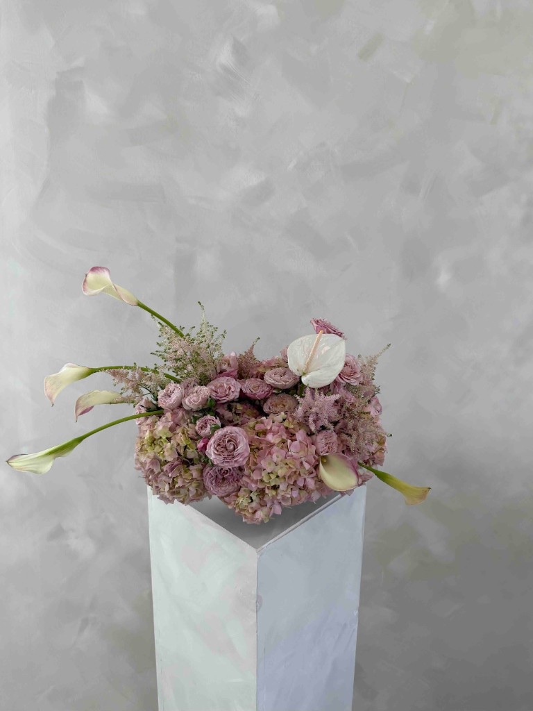 Pastel arrangement with pink and green hydrangeas, lavender roses, pale calla lilies, and a white anthurium in a white bowl on a pedestal