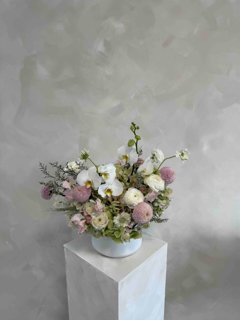 Soft pink, green, and white arrangement with Phalaenopsis orchids, pink pom-pom blooms, white ranunculus, and pale green hydrangeas in a white bowl on a pedestal