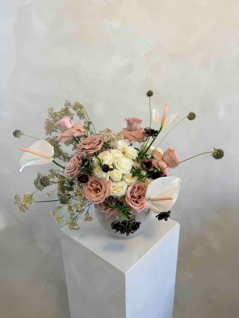 Arrangement of muted pink and cream roses, white anthurium and dark accents in a bowl vase on pedestal