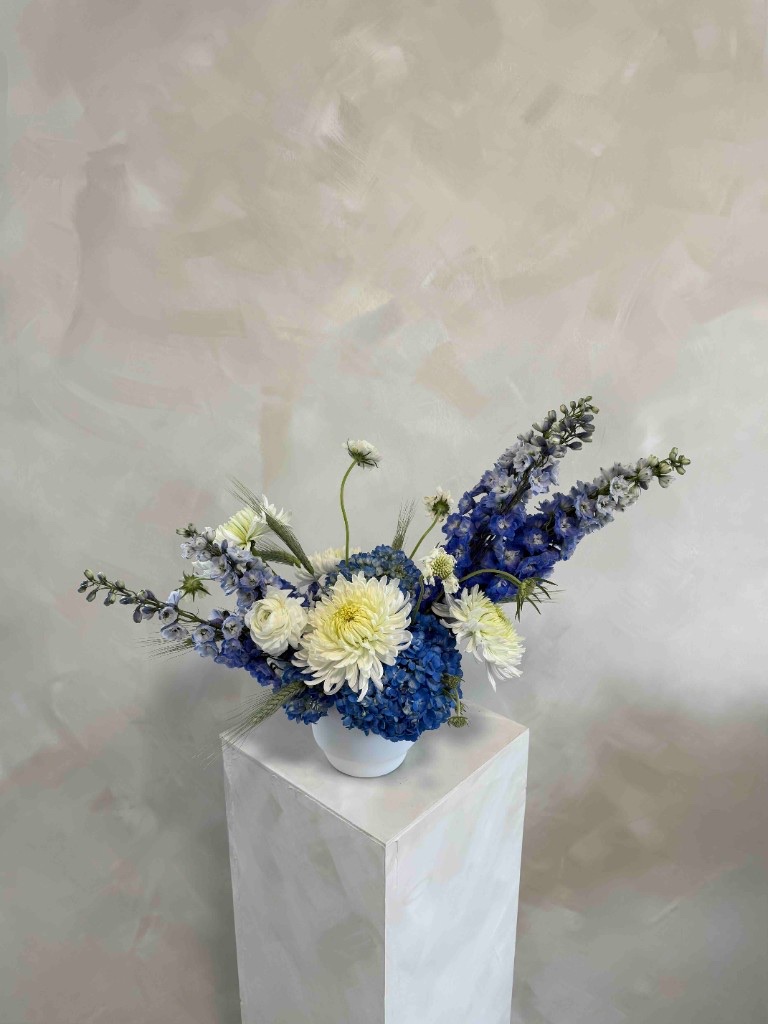 Airy blue, white, and green arrangement with deep blue hydrangeas, a white chrysanthemum, periwinkle delphinium, and white ranunculus in a white bowl on a pedestal