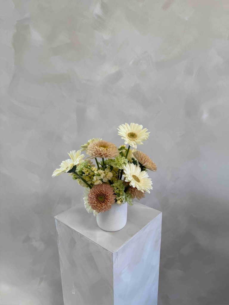 Buttercream and pink arrangement with pale yellow gerbera daisies, dusty peach pompon chrysanthemums, and green filler in a white speckled vase on a pedestal