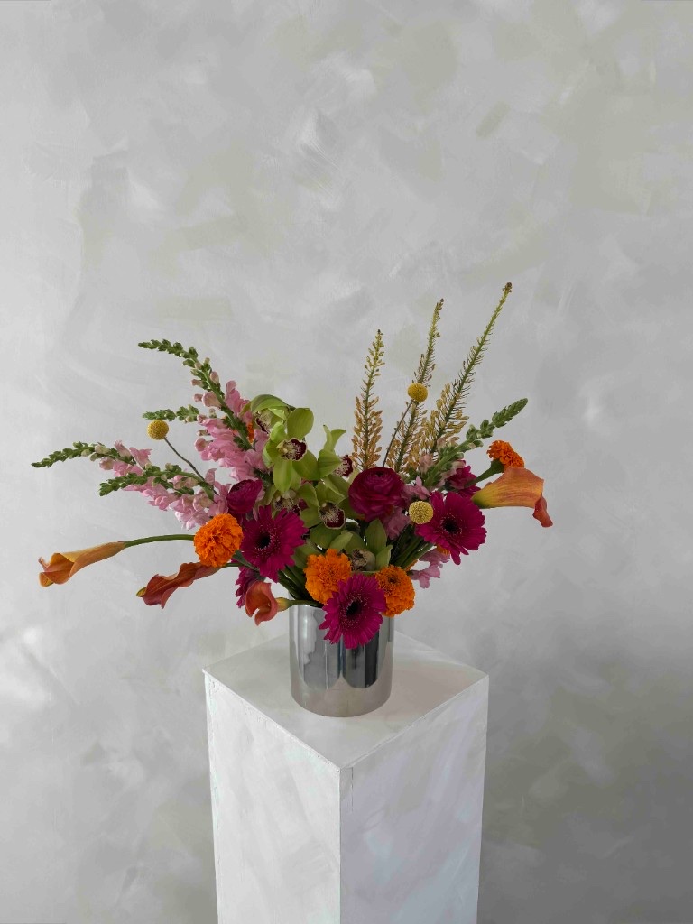 Vibrant summertime arrangement with hot pink gerberas, orange marigolds, lime green orchids, orange calla lilies, and billy balls in a silver vase on a pedestal