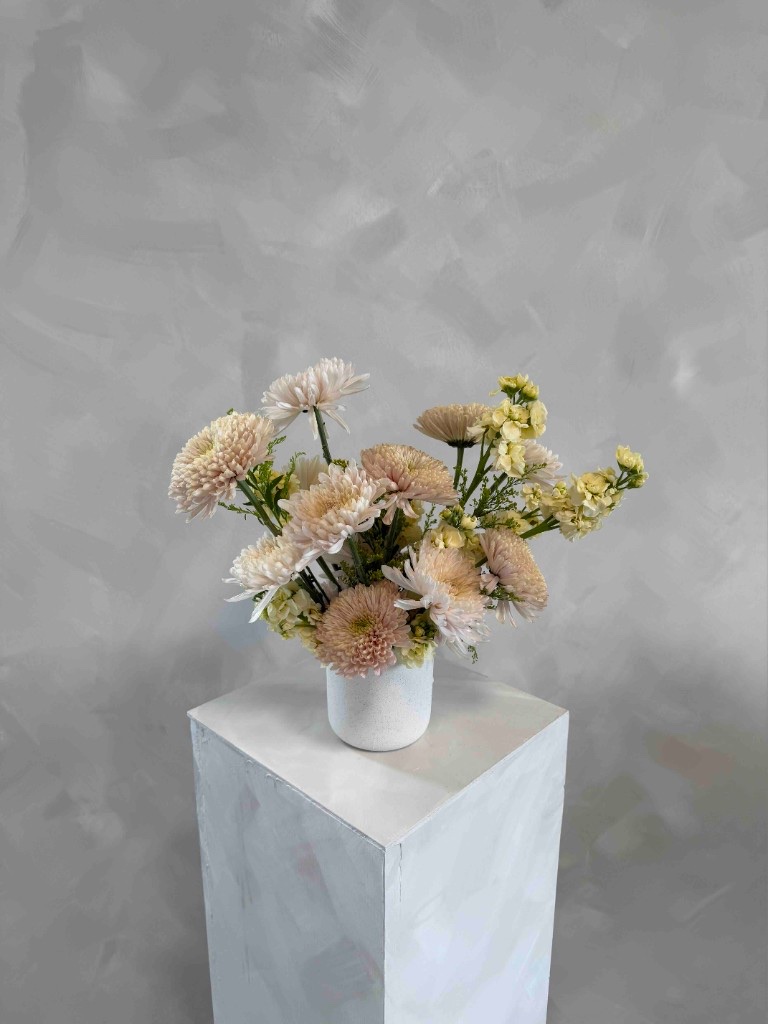 Peach and butter yellow arrangement with dusty pink chrysanthemums and pale yellow stock in a white speckled vase on a pedestal
