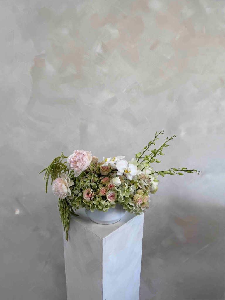 Pastel arrangement with pale pink peonies, light green hydrangeas, spray roses, white orchids, and trailing green amaranthus in a white bowl on a pedestal