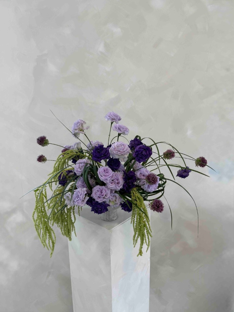 Whimsical purple and green arrangement with lavender and deep purple lisianthus, purple allium, and trailing green amaranthus in a light gray vase on a pedestal