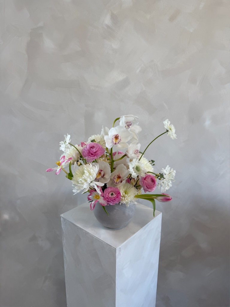 Pink and white arrangement with bright pink ranunculus, white orchids, white chrysanthemums, and lacey filler flowers in a light gray bowl on a pedestal