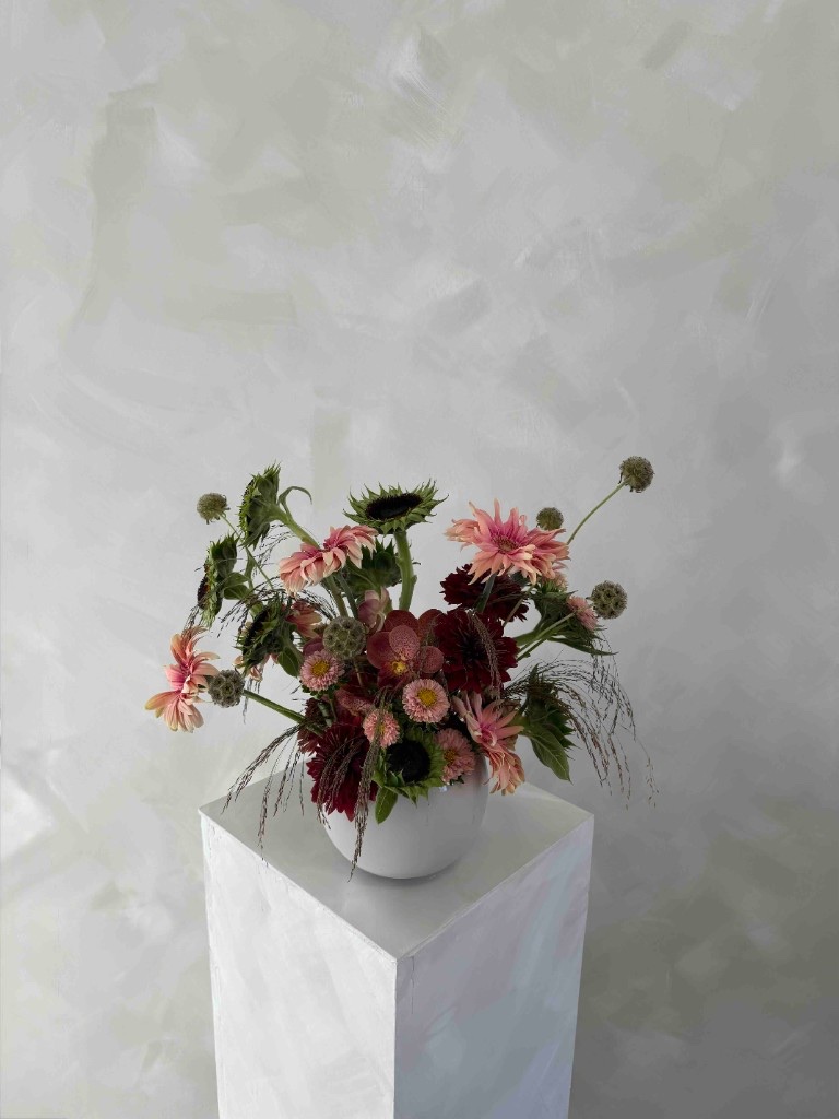 Fall arrangement with pink gerbera daisies, burgundy dahlias, sunflowers, scabiosa pods, and wispy grasses in a white bowl on a pedestal