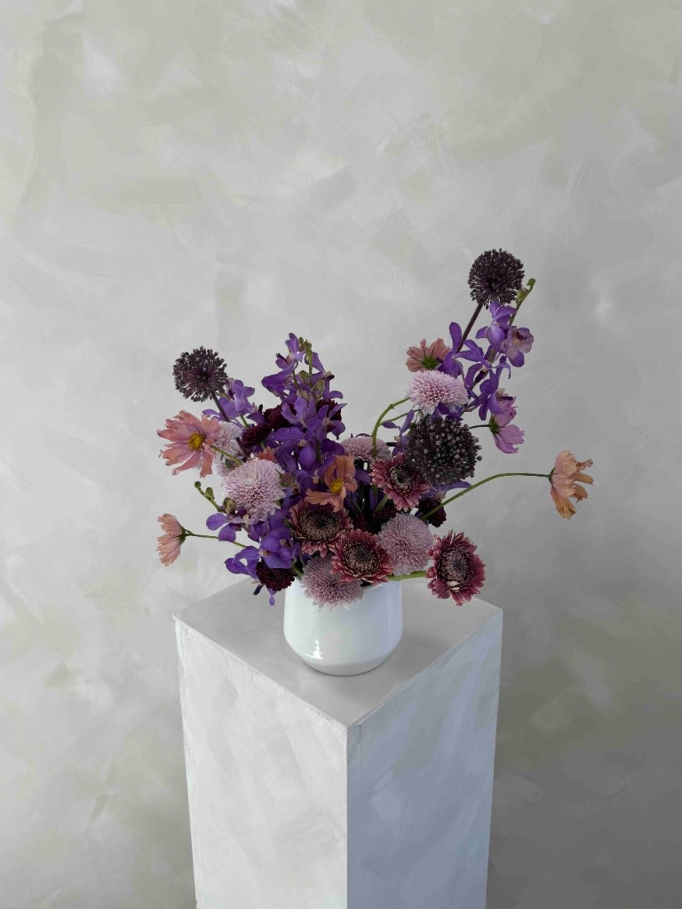 Purple and blush arrangement with delphinium, allium, scabiosa, and chrysanthemums in a white vase on a pedestal against a textured gray wall