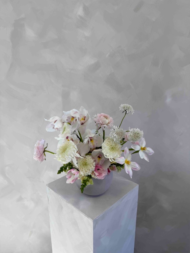 Soft pink and white arrangement with white cymbidium orchids, pink ranunculus, white spider mums, scabiosa, and tulips in a lavender bowl on a pedestal