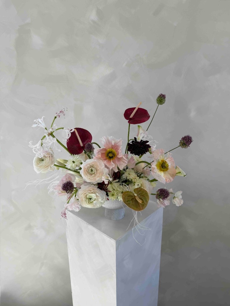 Airy arrangement with burgundy anthurium, blush Iceland poppies, white ranunculus, nerine lilies, allium, and chocolate scabiosa in a white vessel on a pedestal