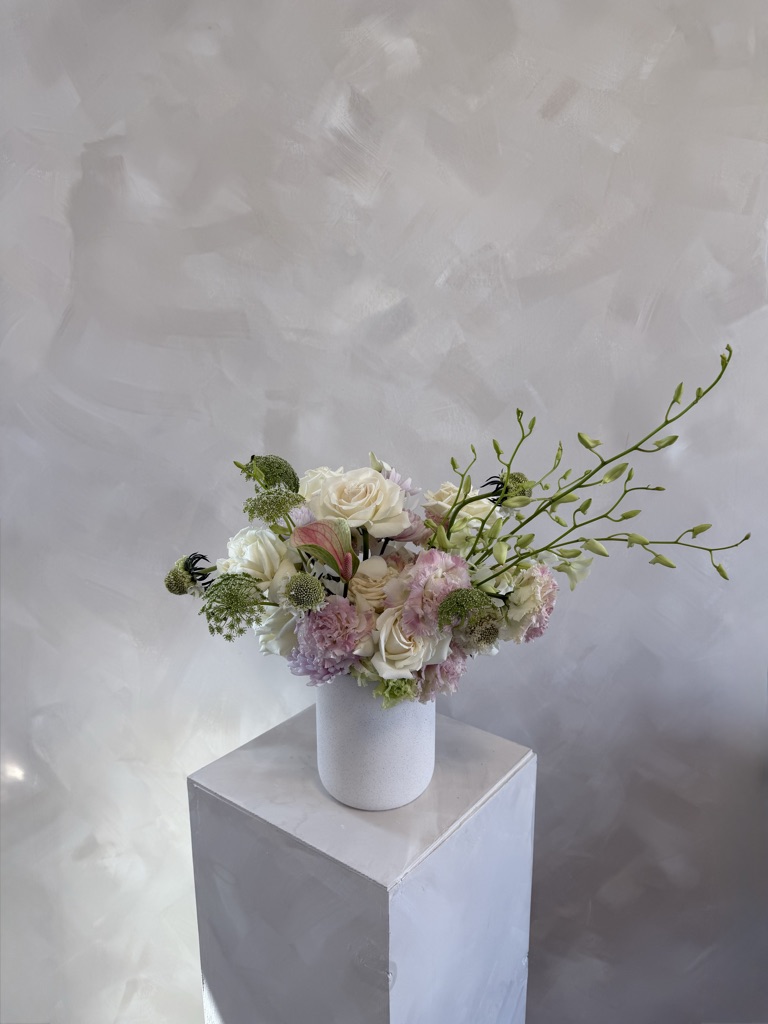 Soft pastel arrangement with white roses, lavender-pink carnations, green trachelium, and arching dendrobium orchid stems in a white speckled vase on a pedestal