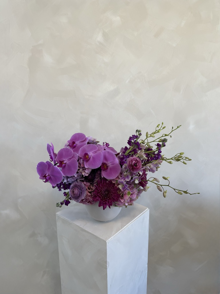 Vibrant purple arrangement with magenta phalaenopsis orchids, lavender ranunculus, burgundy chrysanthemums, hydrangea, stock, and dendrobium orchid stems in a white bowl on a pedestal