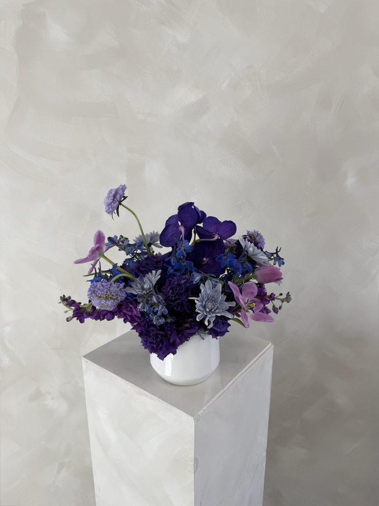 Deep purple and blue arrangement with vanda orchids, purple carnations, delphinium, lavender scabiosa, pink tulips, and dusty blue chrysanthemums in a white vase on a pedestal