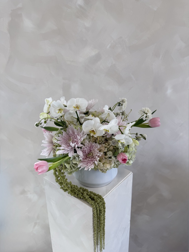 Blush and white arrangement with white phalaenopsis orchids, lavender spider mums, pink tulips, green hydrangea, scabiosa, and cascading green amaranthus in a white bowl on a pedestal